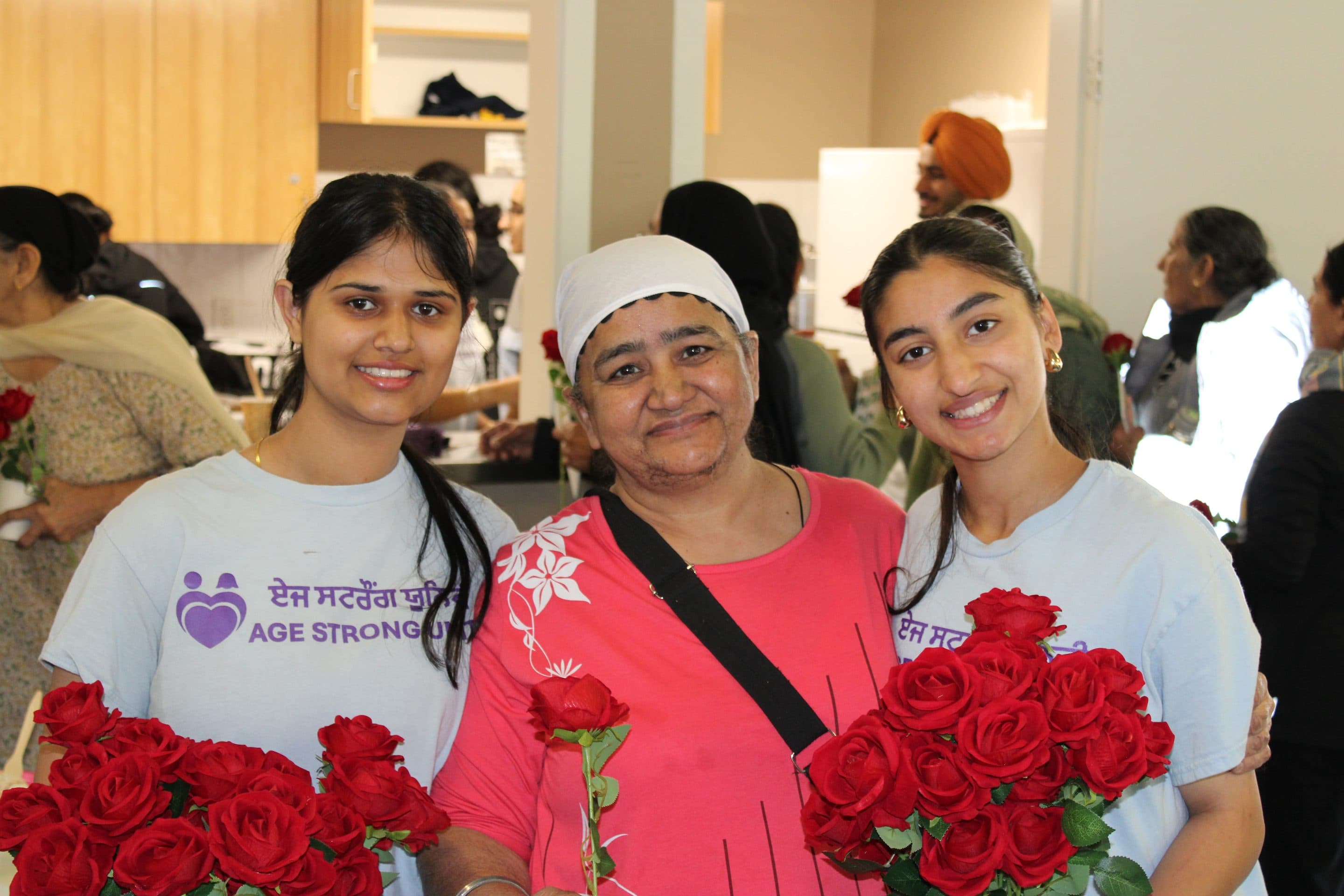 Two volunteers in Age Strong shirts with a matriarch between them, all holding red roses.