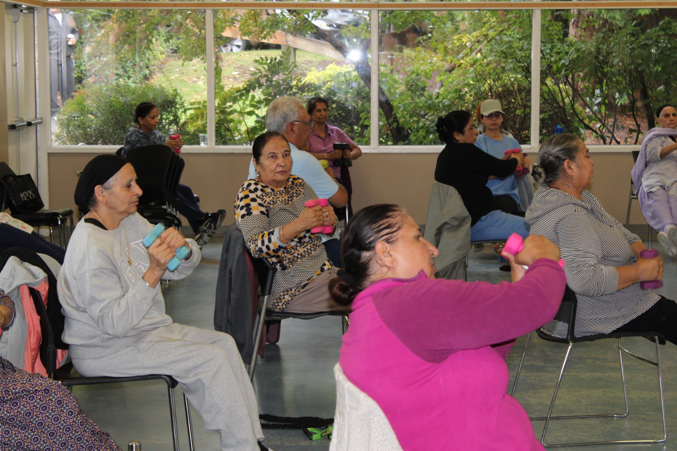 Several women in class lifting small dumbbells, with bright daylight from the windows behind.